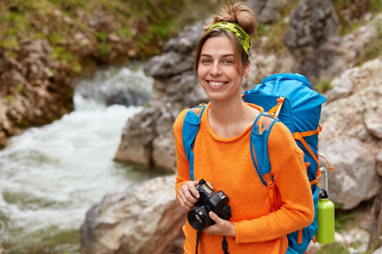 Positive young traveler poses with camera and backpack against canyon, enjoys nature and landscape, nice day, fresh air, poses outdoor with blank copy space. Traveling and recreation concept