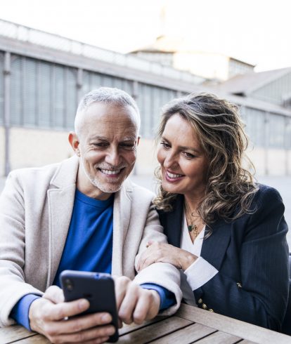 Mid adult couple looking at smartphone while sitting at outdoors table. Carefree senior man and woman using mobile phone together at a terrace in the street.