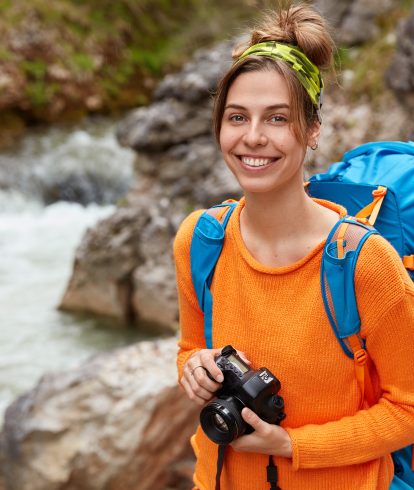 Positive young traveler poses with camera and backpack against canyon, enjoys nature and landscape, nice day, fresh air, poses outdoor with blank copy space. Traveling and recreation concept